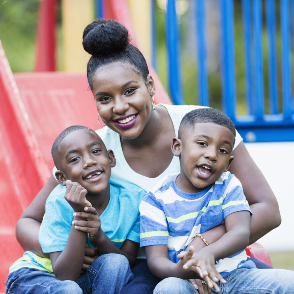 A smiling woman with her hair in a bun sits closely with two young boys on a red slide at a playground. The older boy in a blue shirt holds her arm, while the younger boy in a striped shirt leans against her. They all appear joyful and relaxed.