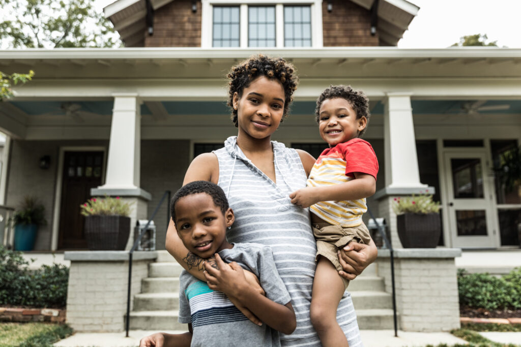 A woman stands in front of a house, smiling and holding a young boy while another boy stands close beside her. All three look happy and are posing for the photo on the porch steps.