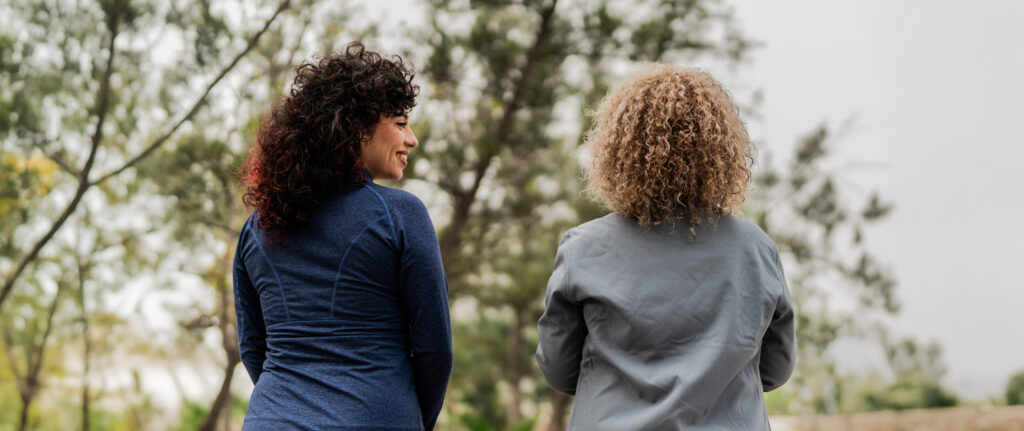 Two people with curly hair, seen from behind, are standing outdoors and facing each other amidst trees on a cloudy day.
