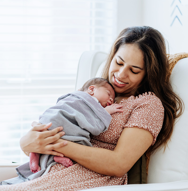 A woman sits in a chair, smiling down at a newborn baby sleeping peacefully on her chest. The baby is wrapped in a striped blanket, and soft natural light filters through the window behind them.