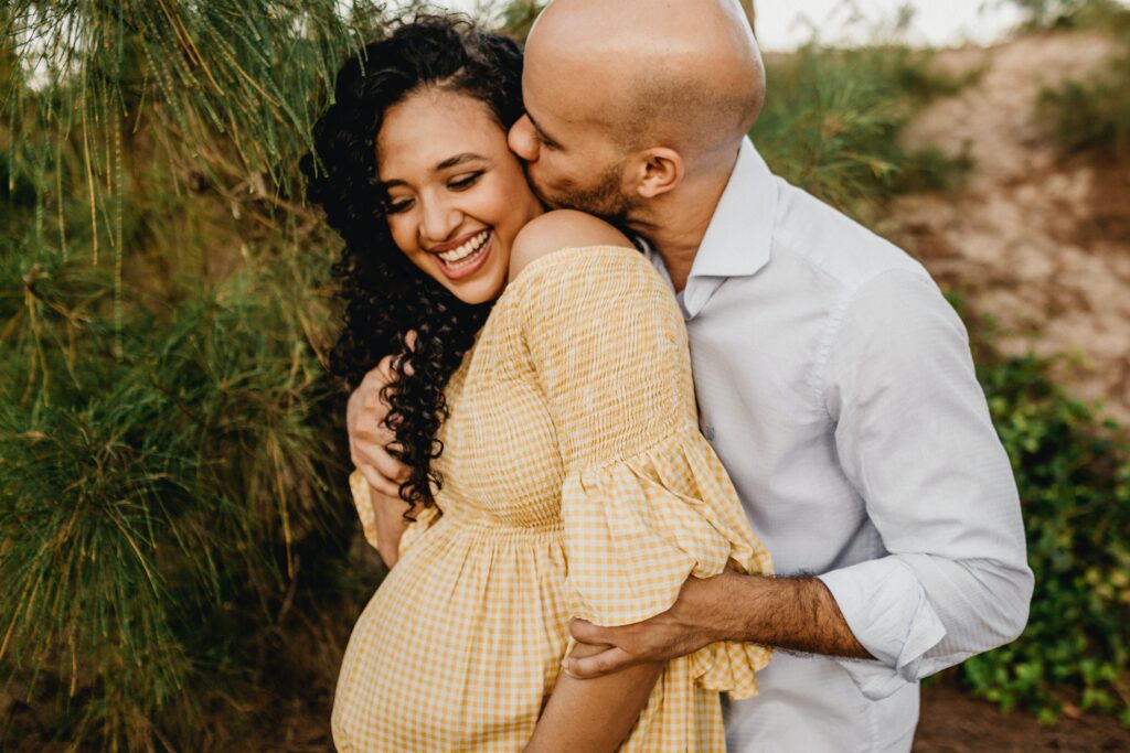 A couple is standing outdoors, surrounded by greenery. The man, wearing a light shirt, embraces and kisses the woman, who is smiling and wearing a yellow checkered dress. The setting appears peaceful and natural.