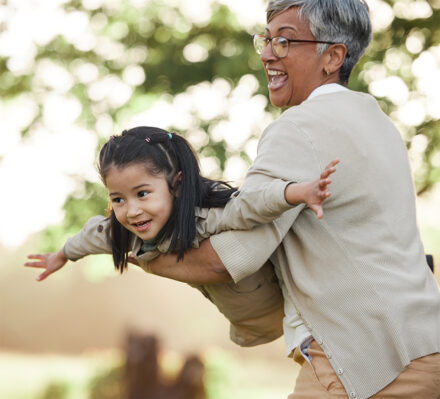 An older adult joyfully lifts a smiling young girl, who stretches her arms out like an airplane, while they play together outdoors in a sunlit, green setting.