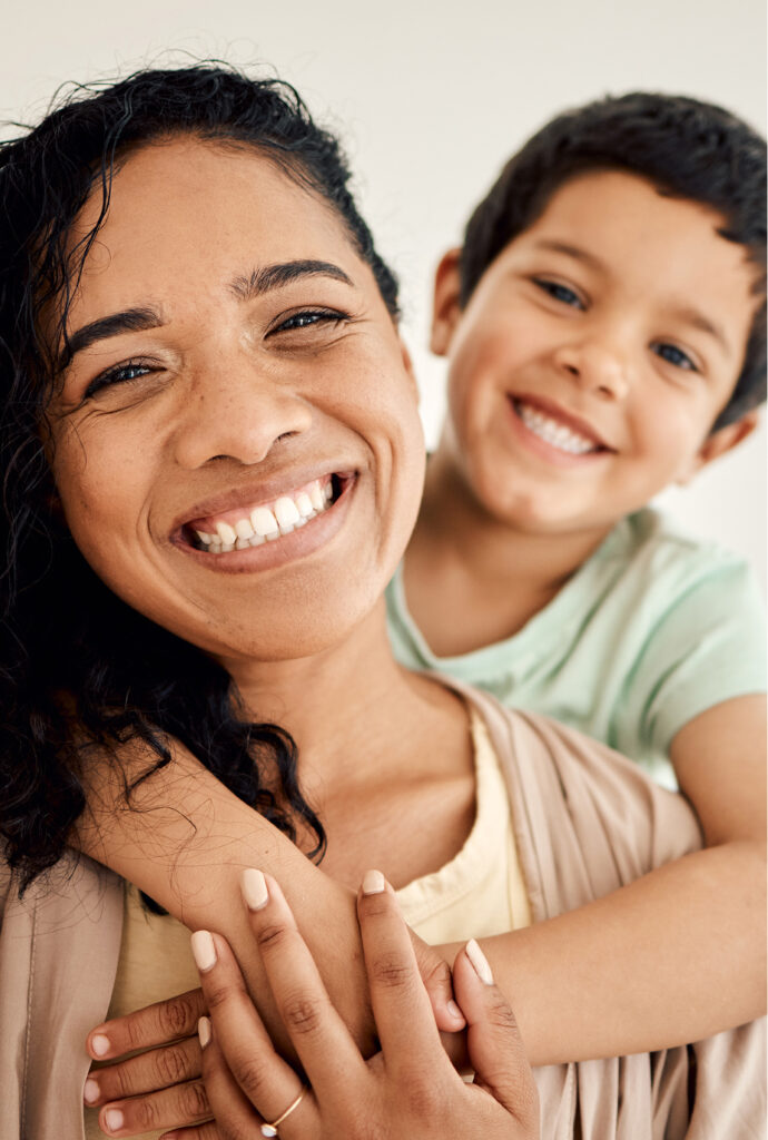 A smiling woman with curly hair stands in front, while a cheerful young boy in a light green shirt hugs her from behind. Both are looking at the camera with happy expressions.