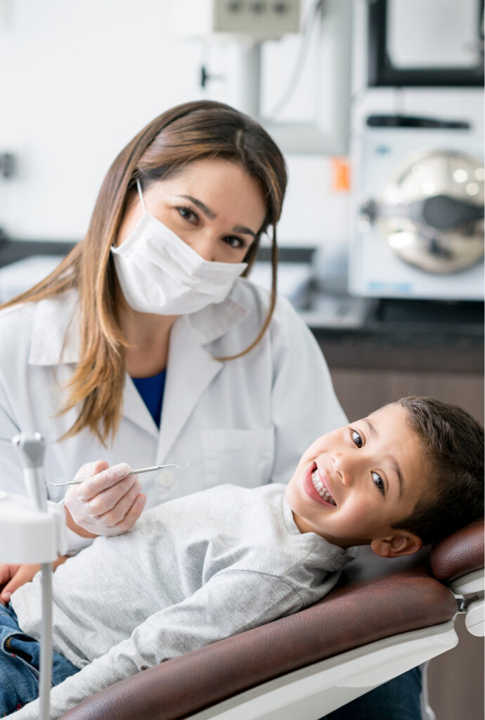 A dentist wearing a mask and white coat is holding dental tools while a young boy smiles and sits in a dental chair during a check-up.