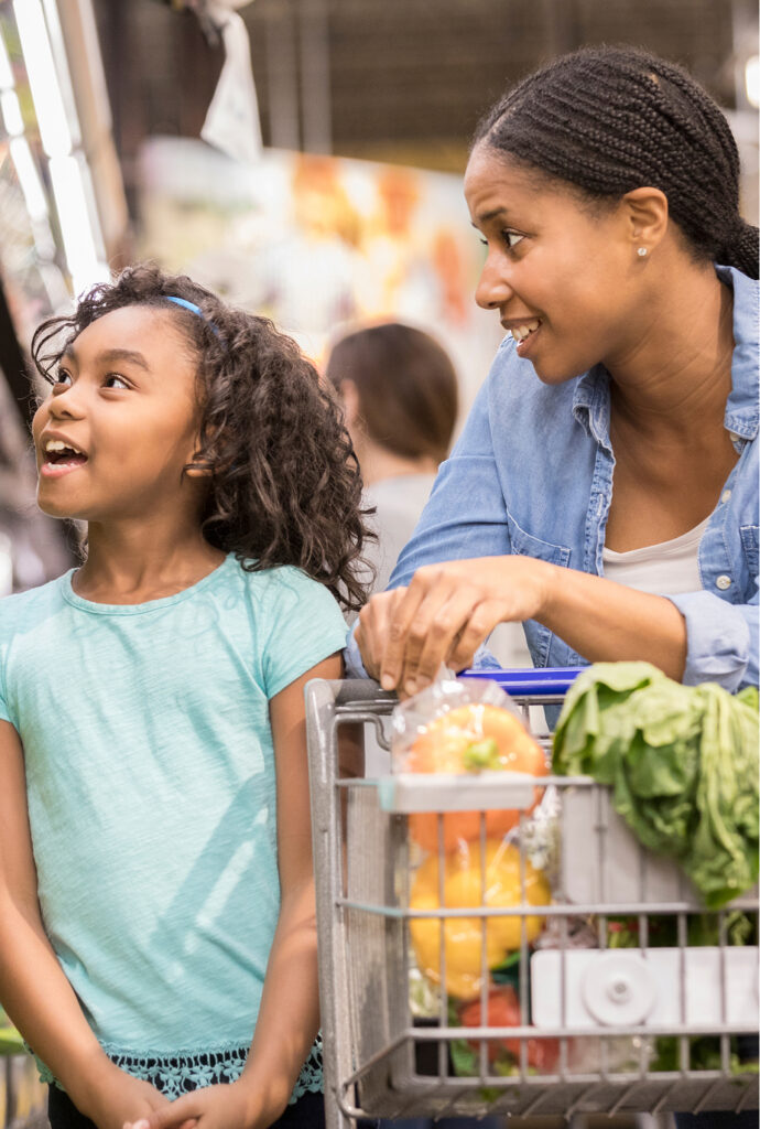 A woman and a young girl smile while shopping in a grocery store. The girl wears a light blue shirt, and the shopping cart is filled with various fruits and vegetables.