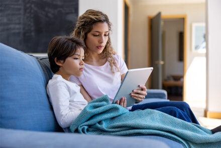 A woman and a child sit on a couch, both looking at a tablet. The child is wrapped in a blue blanket, and the woman is holding the tablet, possibly reading or watching something together.
